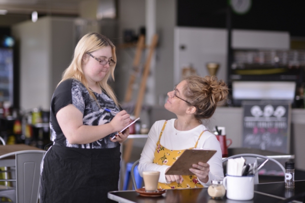 This is an image of a women who is taking an order at a cafe. She is holding a pen and pad. She has longe blonde hair and fair skin, she is wearing a black shirt and an apron. There is a lady seated giving her an order, she is pointing at a menu. That lady has her hair up in a bun. She is wearing a yellow summer dress. 
