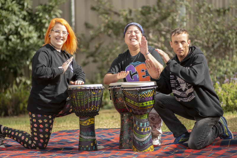 This is a picture of three people playing bongo drums in a local park. There are two women and one man who are all in their mid twenties. They are all smiling and having fun. The first woman has bright orange hair and glasses and tattoos. The second has dark hair and is wearing a colourful headband. The man at the end has dark hair.