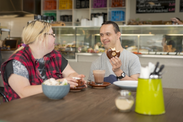 This is an image of two people enjoying a meal at a cafe. There is a man and a woman sitting together on a wooden table. Both have coffee. The man is holding a donut, the women hjas her hands around her coffee glass. 